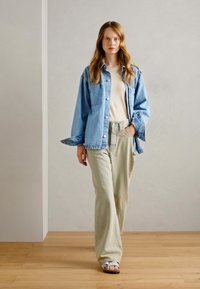 Svijetloplavi traper jakna oversized kroja, krema majica i široke bež hlače. Model nosi svijetle sandale; neutralna pozadina s drvenim podom.