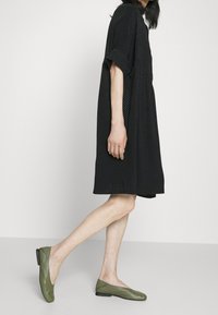 Czarna, teksturowana sukienka z krótkim rękawem i luźnym krojem, w połączeniu z zielonymi mokasynami z niskim obcasem i zaokrąglonym noskiem.