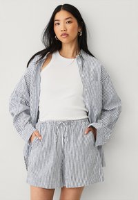 Chemise oversize rayée bleue et blanche et shorts assortis, fabriqués en tissu léger. La chemise a un col, une fermeture à boutons et des poches.