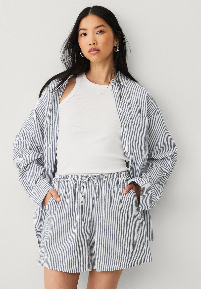 Chemise oversize rayée bleue et blanche et shorts assortis, fabriqués en tissu léger. La chemise a un col, une fermeture à boutons et des poches.