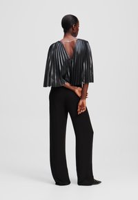 Black pleated cape-style top with metallic sheen, V-back, worn over black wide-leg trousers, showcasing textured fabric and draped silhouette.