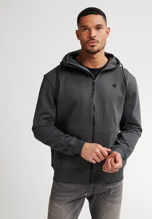 Haut de hoodie zippé gris foncé avec poches avant, capuche réglable et poignets en tricot. Présente un petit logo sur la poitrine et une texture de tissu lisse.