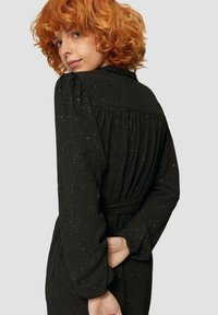 Schwarzes Kleid aus strukturiertem Stoff, mit einem gefalteten Rücken, langen Ärmeln und einem Bindegürtel, akzentuiert mit weißen Sprenkeln.