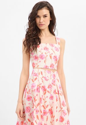 Frau trägt ein ärmelloses, bauchfreies Oberteil und einen hoch taillierten Rock mit rosa und orangefarbenen Blumenmustern auf hellem Hintergrund.