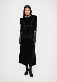 Zwarte fluwelen jurk met korte mouwen, kantdetails en een aansluitende corsage; gecombineerd met lange zwarte handschoenen en puntige hakken.