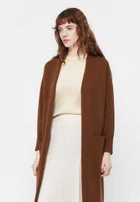 Cardigan lungo marrone in tessuto lavorato a maglia, con polsini a costine e apertura frontale, abbinato a una maglietta beige chiaro e una gonna chiara a tessuto testurizzato.