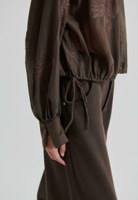 Blouse marron avec broderie florale, taille froncée et manches longues bouffantes ; associée à un pantalon ample et texturé de couleur marron.