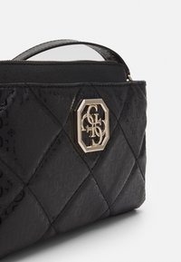 Sort quiltet håndtaske med subtilt monogrammønster og guldfarvet oktagonal logo-plade på forsiden, delvis lynlås og håndtag synligt.