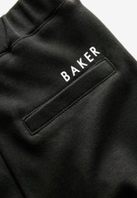 Čierne bavlnené joggery s textúrovaným povrchom, s obdĺžnikovou zadnou vreckom a nápisom "BAKER" v bielej farbe. Jednoduchý, funkčný dizajn.