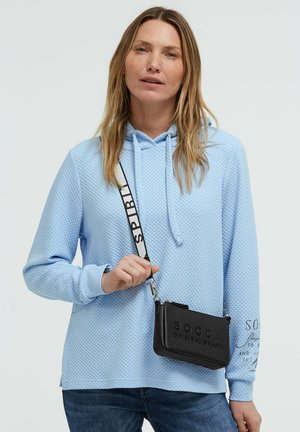 Woman wearing a light blue textured hoodie and jeans, holding a black crossbody bag with a printed strap over her shoulder.