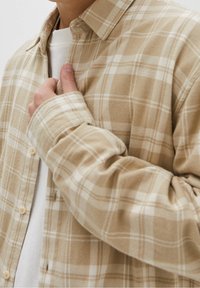 Persona con camisa de cuadros beige abotonada sobre camiseta blanca, sujetando el cuello con la mano izquierda.