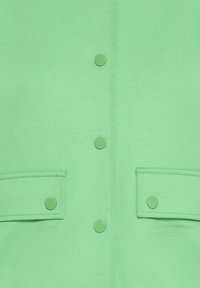 Zelena srajca z gladko teksturo, ki ima pet okroglih gumbov in dva dodatna žepa. Preprost dizajn in enotna barva po celotnem izdelku.