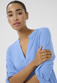 Blouse texturée bleu clair avec un col en V, des manches repliées et une coupe ample. Le tissu présente un motif subtil et une finition lisse.