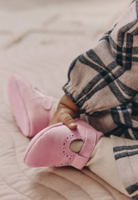 Scarpe morbide in pelle rosa pallido con punte arrotondate, dettagli traforati e cinturino laterale. Mano posizionata sopra la scarpa, su una superficie trapuntata.