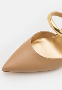 Chaussure plate en cuir beige à bout pointu avec doublure intérieure métallisée dorée et boucle de bride de cheville dorée brillante.