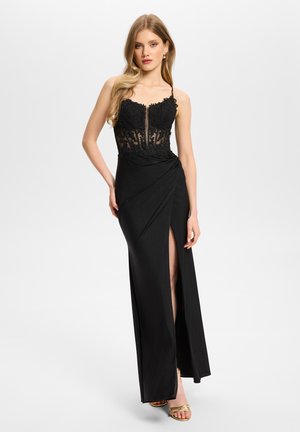Jeune femme aux longs cheveux blonds portant une robe de soirée noire à corsage en dentelle avec une fente haute sur la cuisse et des talons ouverts dorés.