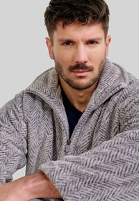 Forro polar gris en patrón de espiga, tela texturizada, mangas amplias, cuello alto, usado sobre una camisa azul marino oscura. Primer plano del torso superior del modelo.