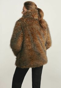 Brązowy płaszcz z sztucznego futra o puszystej strukturze, z wysokim kołnierzem i oversize'owym krojem, stylizowany z czarnymi spodniami.