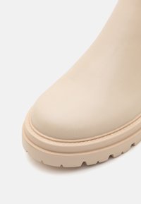 Beige nahkasaapas nilkkamallilla, pyöreällä kärjellä, pintakuvioinnilla ja paksulla, vaalealla pohjalla, jossa on uramalli.