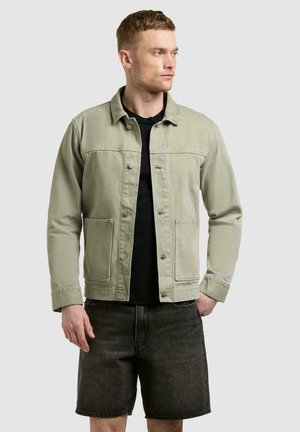 Man draagt een lichtgroene jas met knopen over een zwart shirt en donkere denim shorts, kijkt naar rechts tegen een effen achtergrond.