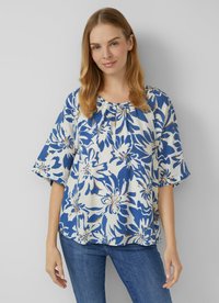 Frau in einer lockeren weißen Bluse mit großem blauen und beigen Blumenmuster und Dreiviertelärmeln, kombiniert mit blauen Jeans, vor einer weißen Wand.