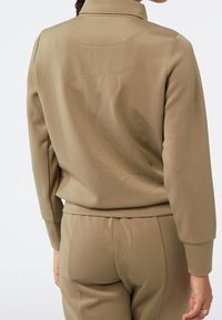 Tan atletisk zip-up jacka gjord av slät tyg, med ståkrage, ribbade mudd och figurnära design. Bakvy visar sömdetaljer.