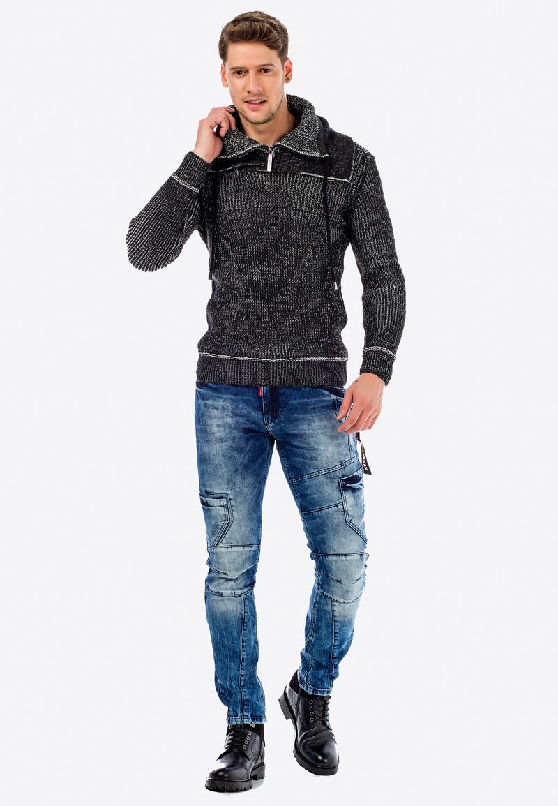 Zwart en grijs gebreid sweater met hoge nek en rits; gecombineerd met verouderde blauwe jeans met cargozakken; gedragen met zwarte laarzen.