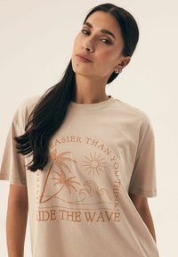 Beżowa koszulka bawełniana z graficznym wzorem przedstawiającym palmy, słońce i fale. Na tekście widnieje napis "Może łatwiejsze, niż myślisz, złap falę."
