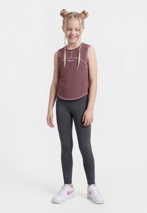 Hymyilevä tyttö, jolla on palmikkoponnarit ja ruskea hihaton Nike Pro -toppi, tummanharmaat legginsit ja valkoiset lenkkarit, joissa on vaaleanpunaiset yksityiskohdat.