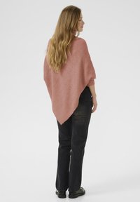 Poncho en tricot rose avec un motif texturé, ayant une coupe ample et un ourlet arrondi, associé à un jean noir et des chaussures noires.