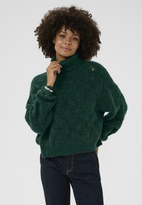 Per didelis žalias megztinis su aukštu apykakle, tekstūruotu raštu, nukritusiomis petnešomis ir gumbuotomis rankovėmis. Derinamas su tamsiomis džinsomis.