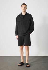 Chemise griffée noire surdimensionnée avec deux poches poitrine, portée avec un short noir et des sandales noires, sur un mannequin devant un fond uni.