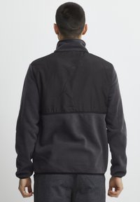 Pull en polaire avec une partie supérieure en nylon noir. Couleur gris foncé, col montant et poignets élastiques. Matériau en polaire texturé.