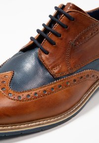 Scarpa oxford in pelle marrone con pannello testurizzato blu navy, dettagli brogue e lacci scuri. Suola cucita con bordo chiaro a contrasto.