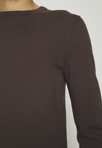 Pull en maille brun foncé à manches longues avec un col rond. Présente une texture côtelée et une silhouette ajustée. Détails de coutures sur les épaules.