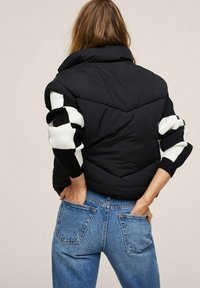 Schwarze Steppweste mit hohem Kragen, ausgestattet mit gestreiften schwarz-weißen Strickärmeln, getragen mit hellblauen Jeans. Strukturierter Stoff und gepolstertes Design.