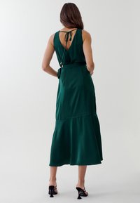 Grön satinklänning med figurnära bodice, öppen rygg med knytning och lager-på-lager kjol. Bärs med svarta klacksandaler.