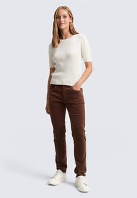 Weißer gerippter Kurzarmpullover mit strukturiertem Design, kombiniert mit brauner Cordhose und weißen Turnschuhen. Das Modell steht selbstbewusst.