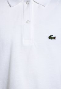 Valkoinen polo-paita, joka on valmistettu teksturoidusta puuvillasta. Paita sisältää kolmen napin kauluksen ja vasemmassa rinnassa on pieni vihreä krokotiililogo brodeerattuna.