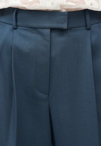 Παντελόνια σε σκούρο μπλε ναυτικό χρώμα, ψηλόμεσα και ραμμένα με ζώνη και πιέτες μπροστά, σε συνδυασμό με λευκή μπλούζα από δαντέλα, φορεμένη από μέσα.