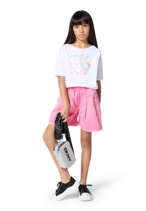 White t-shirt with reflective "DKNY" logo, pink elastic-waist shorts, and black sneakers. She holds a silver crossbody bag with "DKNY" text.