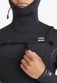 Osoba w czarnym kombinezonie piankowym z kapturem, ściągająca zamek na klatce piersiowej, pokazując logo Billabong na piersi i rękawie.