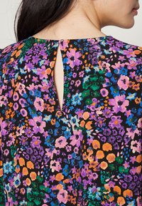 Femme portant une robe fleurie multicolore avec des fleurs violettes, orange, vertes et bleues, présentant une petite ouverture en forme de trou de serrure dans le haut du dos.