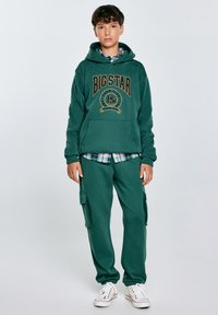 Completo di felpa e pantaloni della tuta verdi, con tasca a marsupio, logo "BIG STAR" e camicia a quadri sotto. Sneakers bianche completano l'outfit.