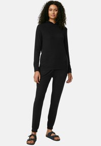 Svart huva sweatshirt och matchande joggers i mjukt tyg, med långa ärmar och ribbade muddar. Bärs med svarta sandaler.