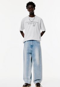 Tricou gri cu mânecă scurtă, inscripționat cu textul "CLOAKROOM SERVICE", asortat cu blugi largi de culoare albastră deschisă și pantofi negri cu șiret.