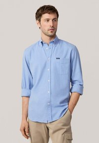 Camisa de botones azul claro con un bolsillo en el pecho, mangas remangadas y una pequeña etiqueta en el lado izquierdo. Hecha de una tela texturizada.