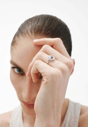 Anillo de plata con una piedra azul ovalada rodeada de pequeñas piedras blancas, con un diseño de banda elegante. Mano posicionada cerca de la cara.