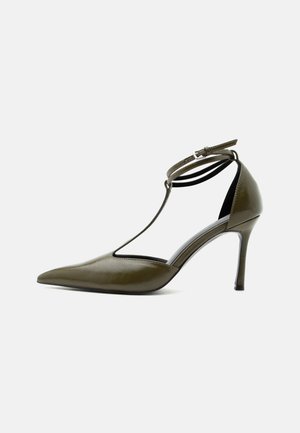 Olivengrøn spids pumps med ankelrem og T-rem design, blank finish, slank stilet hæl, på hvid baggrund.