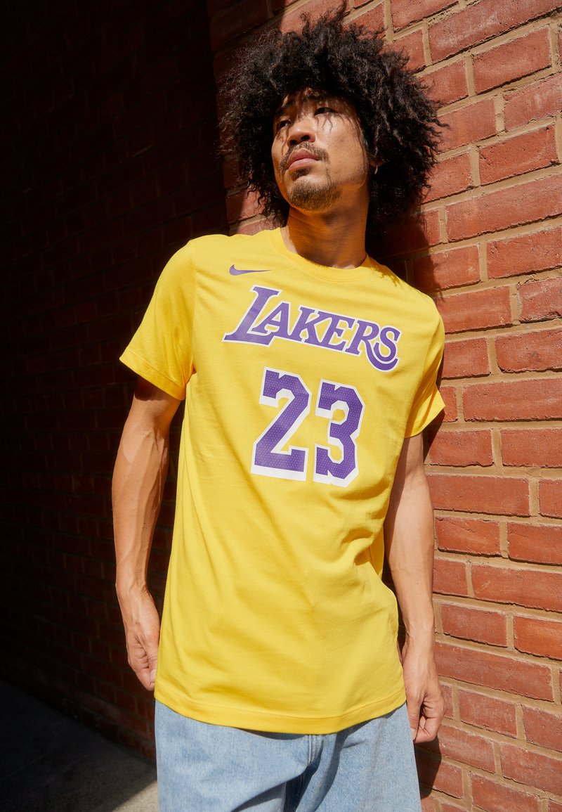 Gul bomulls T-skjorte med "LAKERS" i lilla og nummer "23" trykt i hvitt. Casual passform, korte ermer. Bært med lyseblå jeans.